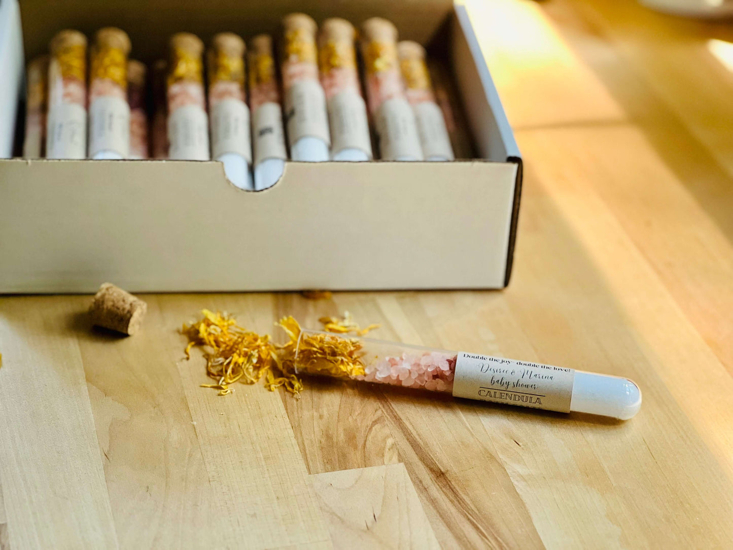 Calendula bath salts in a glass tube with dried petals, displayed on a wooden surface with a box in the background.