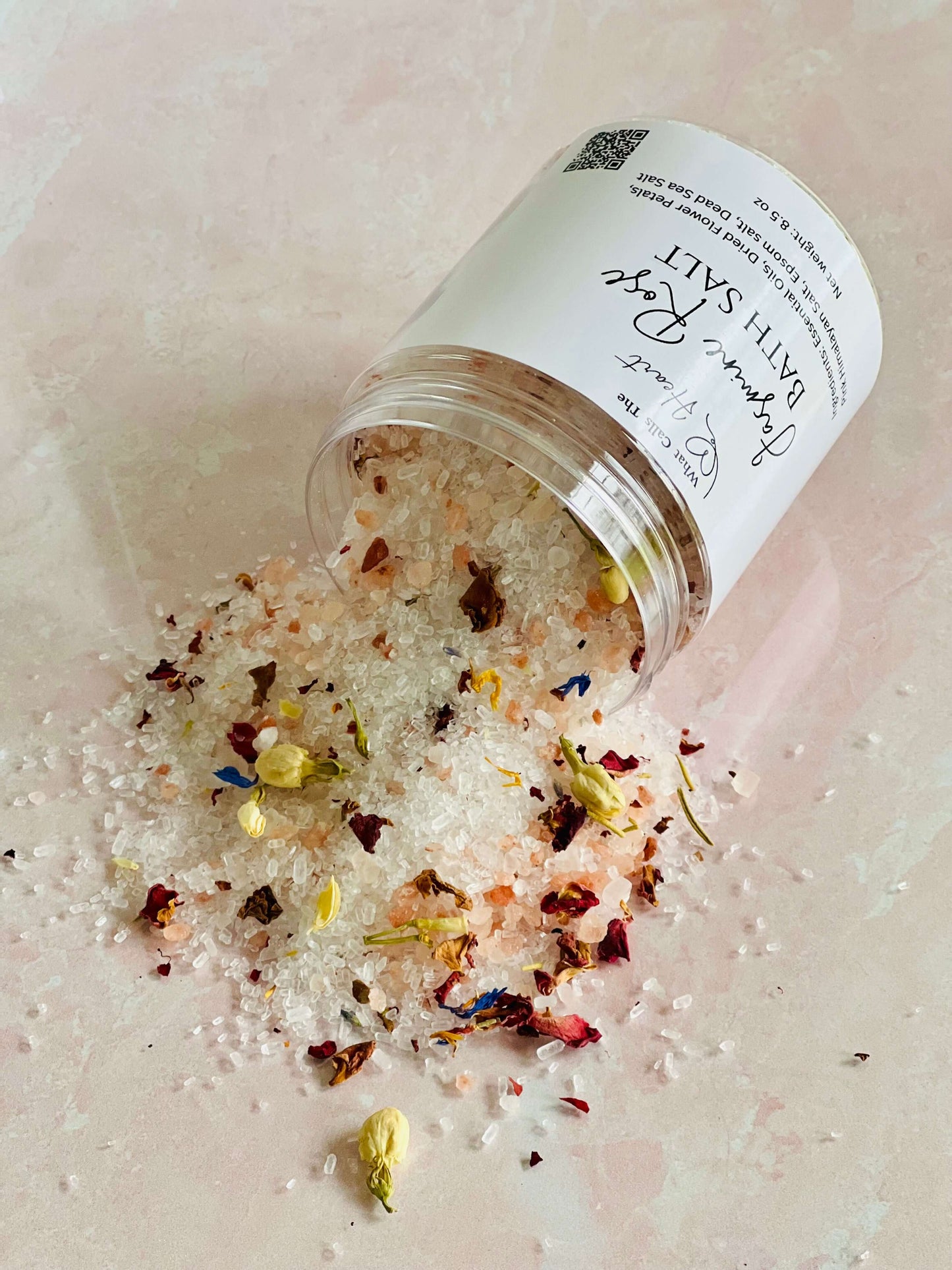 Jasmine Rose Bath Salts spilling from a clear container, featuring flower petals and mineral-rich crystals.