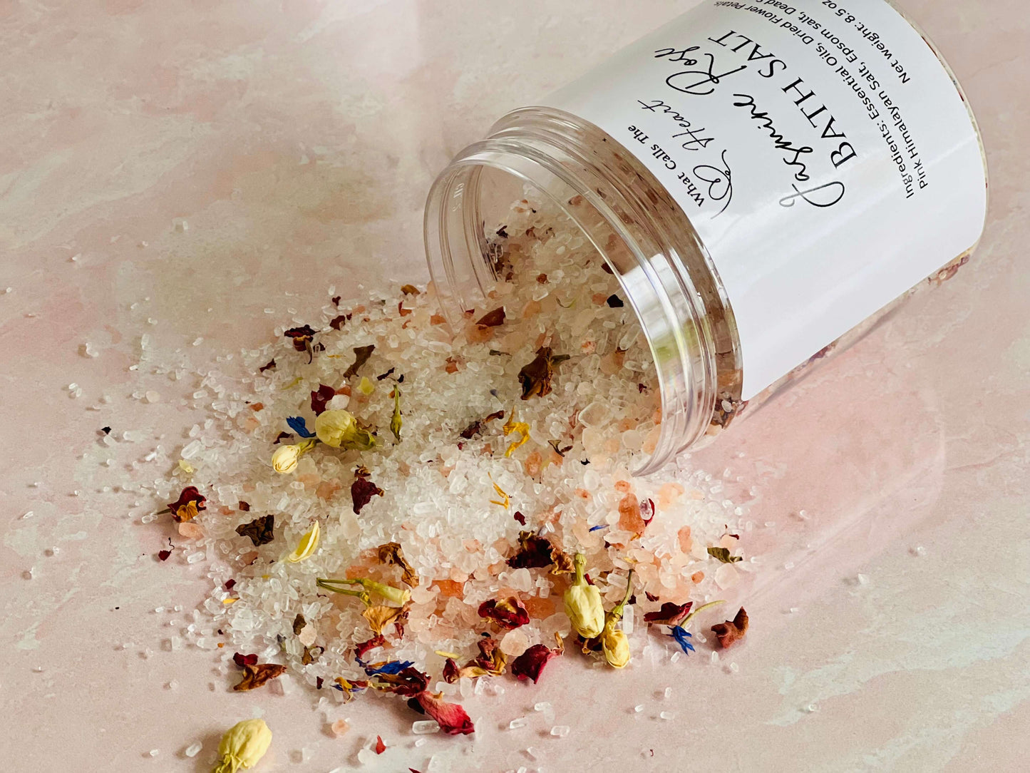Jasmine Rose bath salts spilling from a container with colorful dried flowers and crystals on a pink surface.