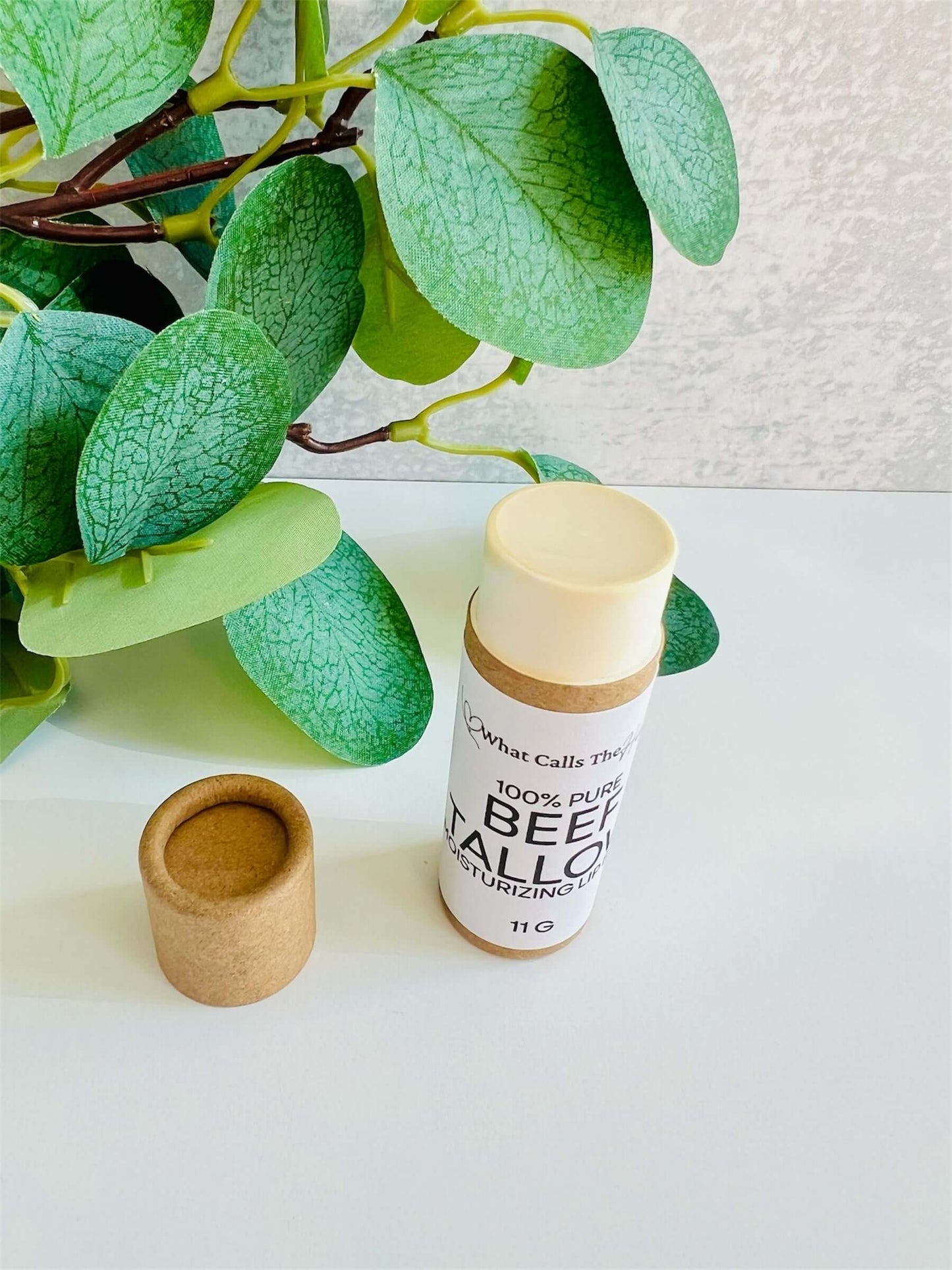 Beef tallow organic lip balm tube next to its lid on a white surface with greenery in the background.