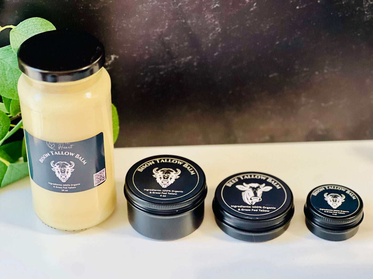 Four sizes of organic bison tallow balm jars displayed with green foliage in the background, promoting skin nourishment.