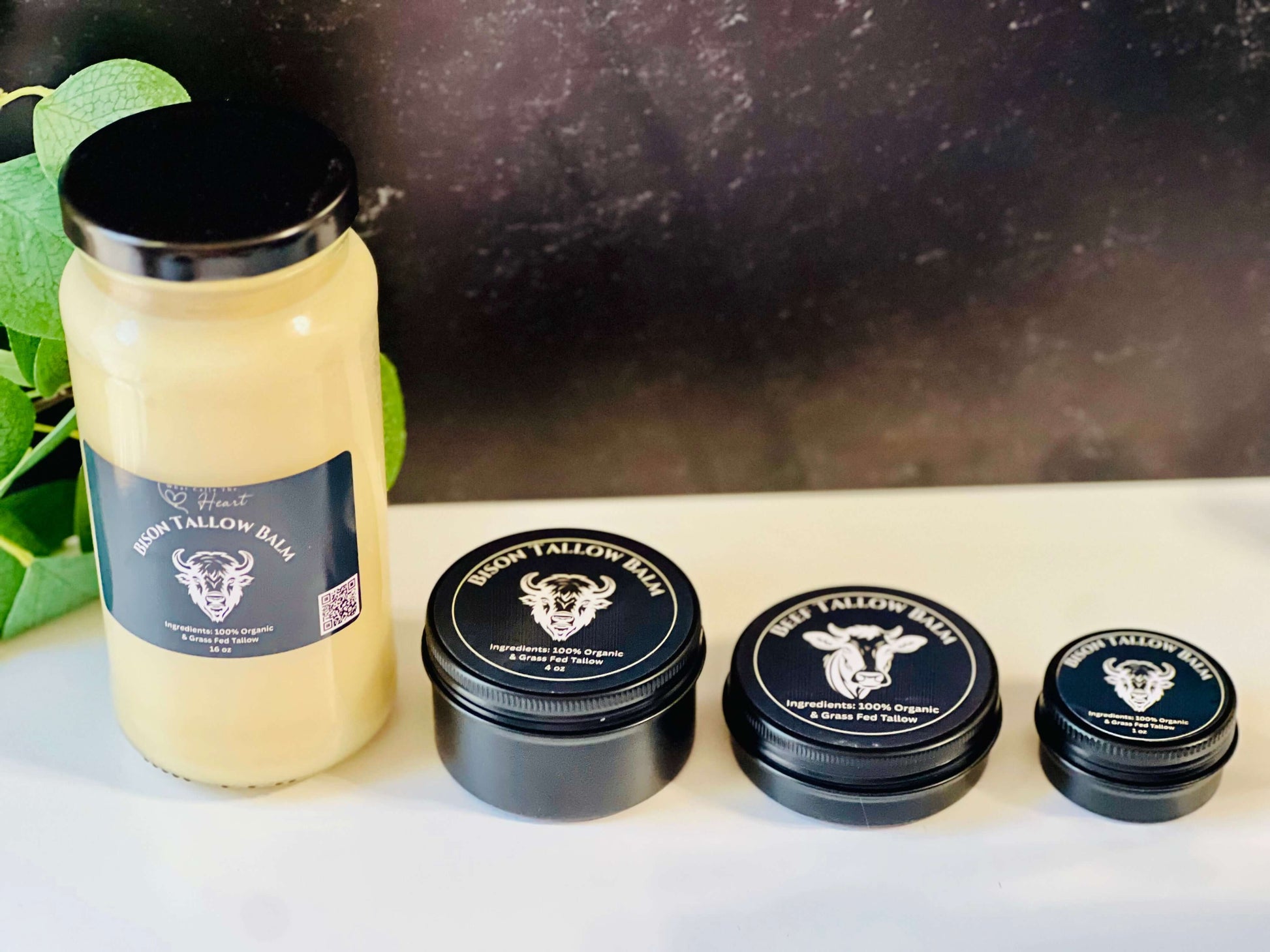 Four sizes of organic bison tallow balm jars displayed with green foliage in the background, promoting skin nourishment.