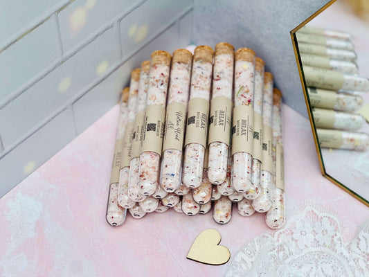 Stack of mixed bath salts in glass tubes with natural ingredients, displayed on a pastel backdrop with a heart accent.
