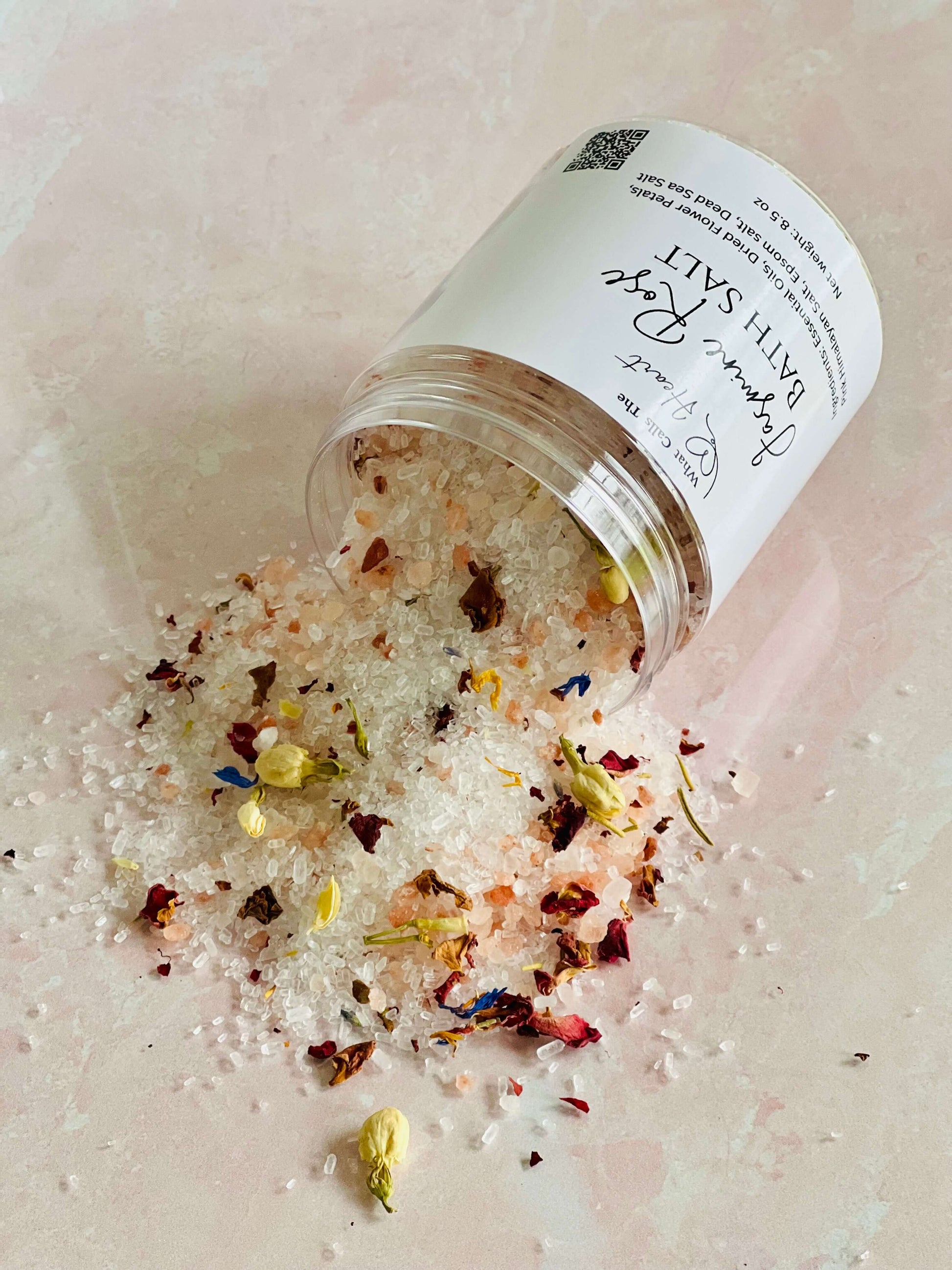 Jasmine Rose Bath Salts spilling from a clear container, featuring flower petals and mineral-rich crystals.