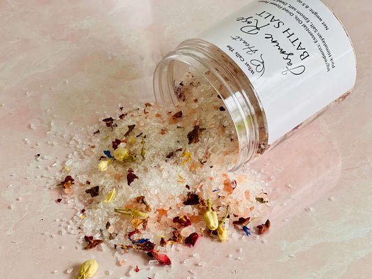 Jasmine Rose bath salts spilling from a container with colorful dried flowers and crystals on a pink surface.