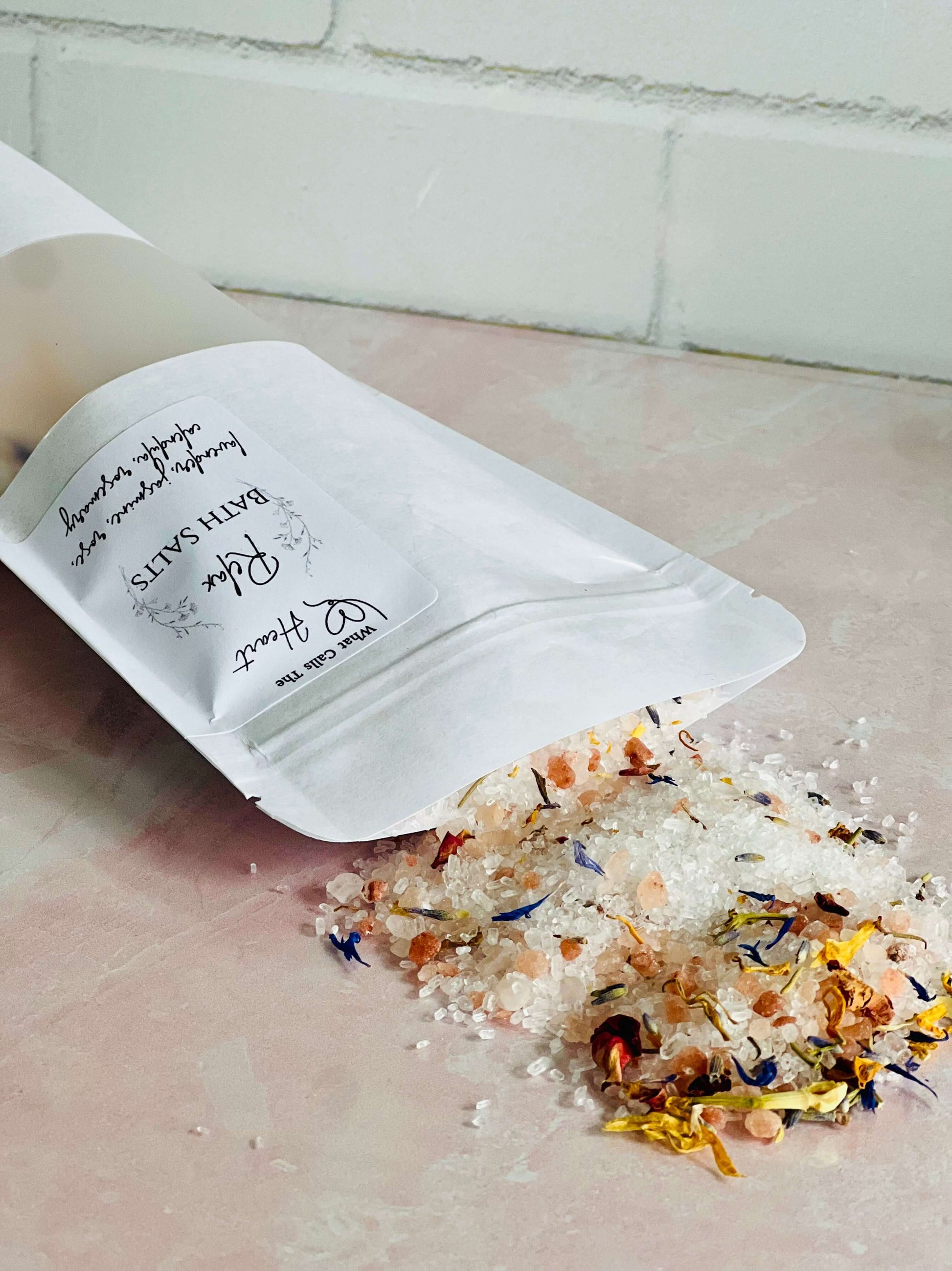 Spilled bath salts with flowers from a biodegradable pouch on a pink surface, promoting relaxation and natural ingredients.